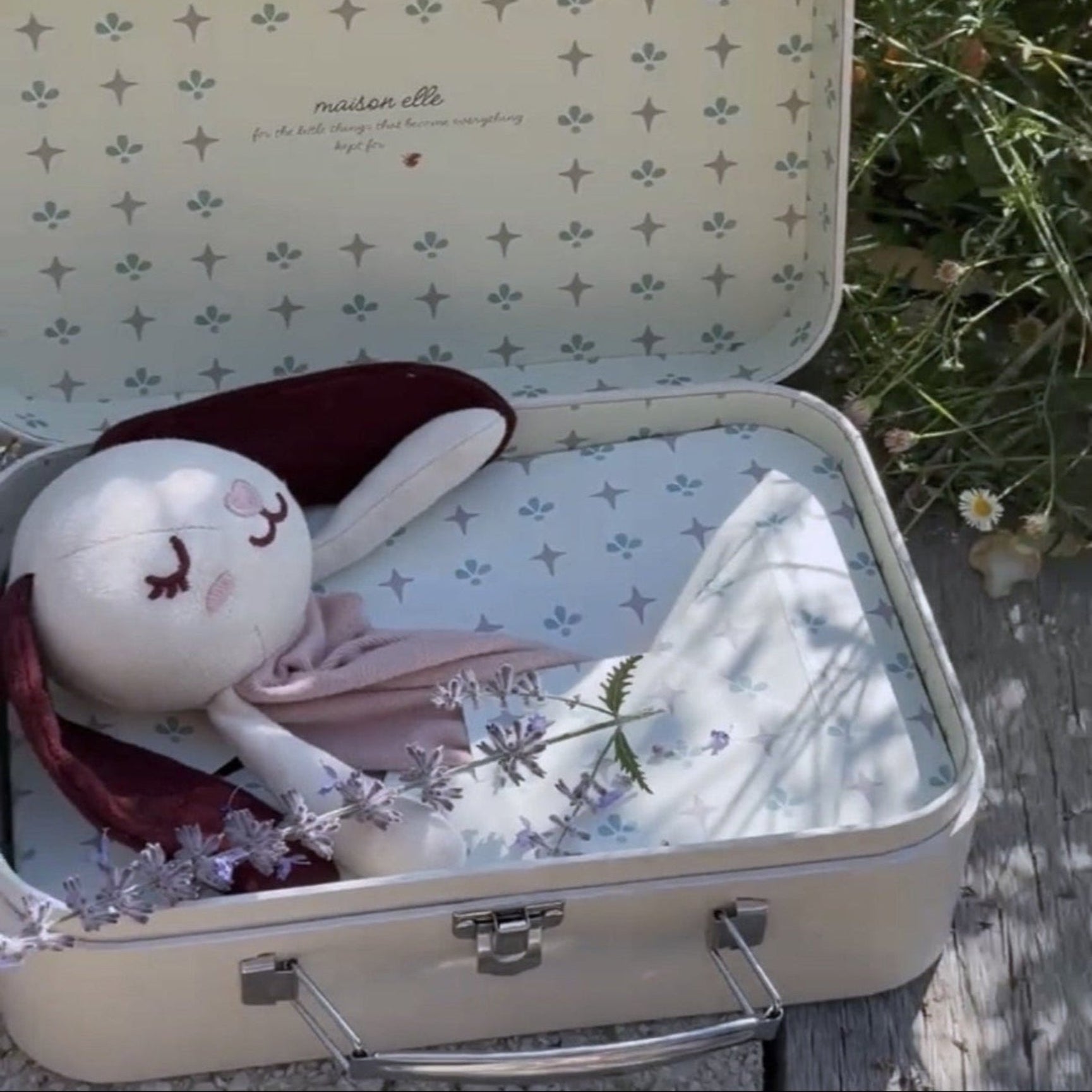 A cream-colored vintage suitcase sits open on a wooden surface outdoors, surrounded by green grass and wildflowers. Inside the suitcase is a white plush bunny doll wearing a burgundy top and light pink skirt, styled with small white flowers.