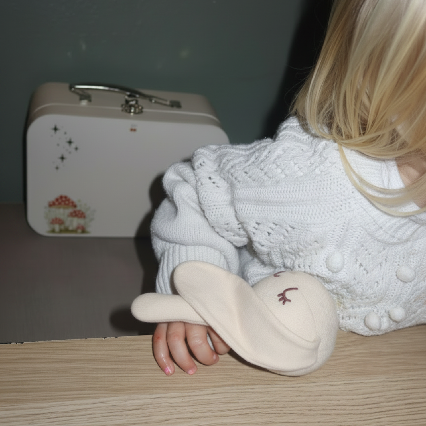 A child in a white knit sweater holds a sleepy softie while sitting at a wooden table, with a white keepsafe box in the background.