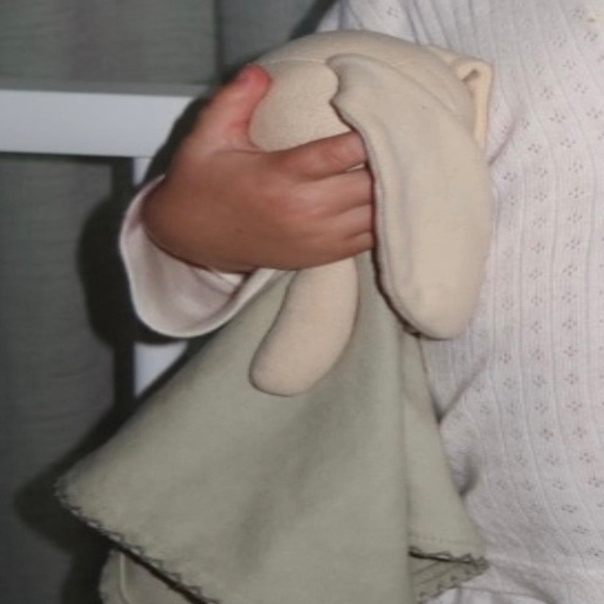 A close-up of a person holding a soft, light beige sleepy softie, and the background is a dark gray curtain.