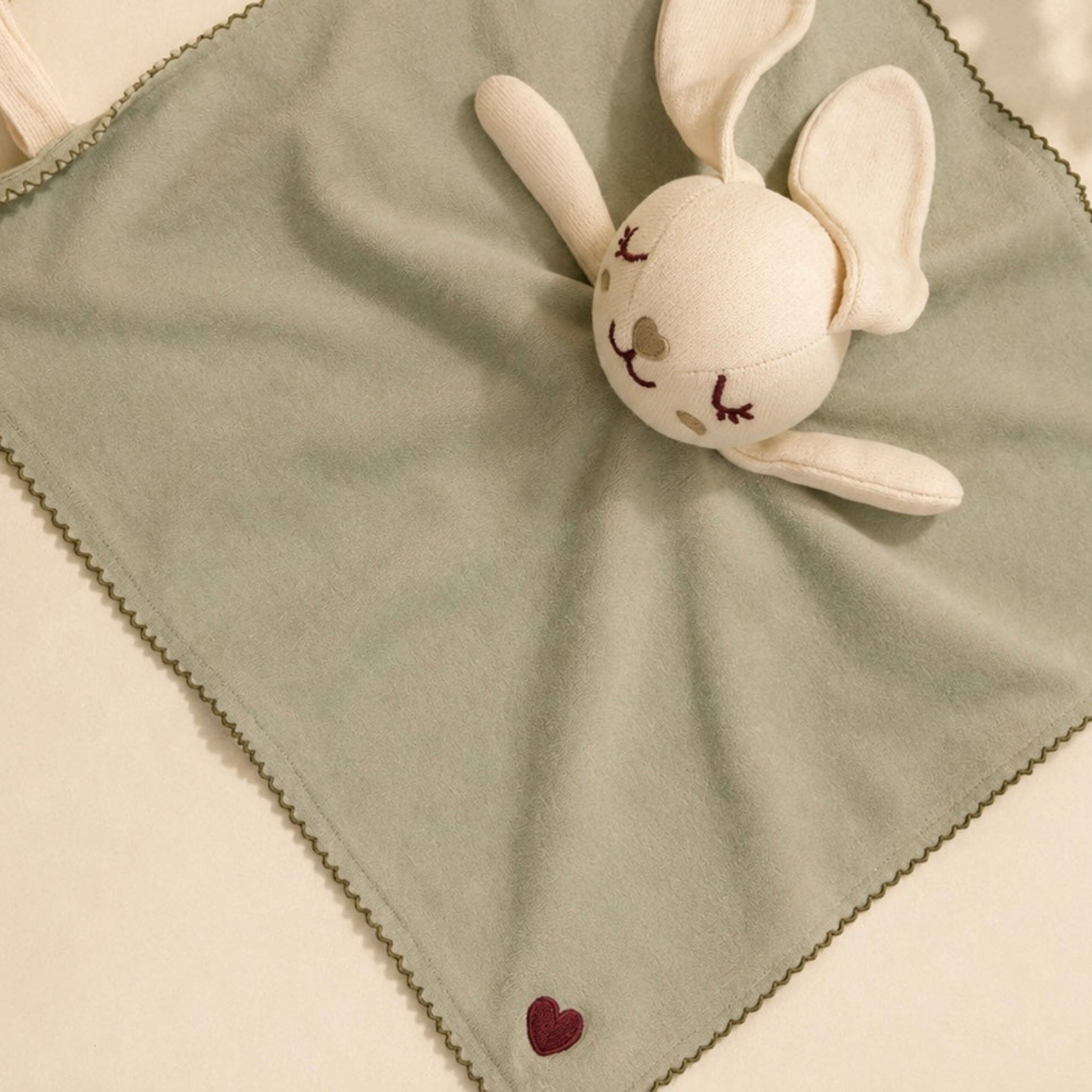 A sage green baby security blanket with a plush white bunny attachment, featuring a small embroidered red heart near the bottom corner.