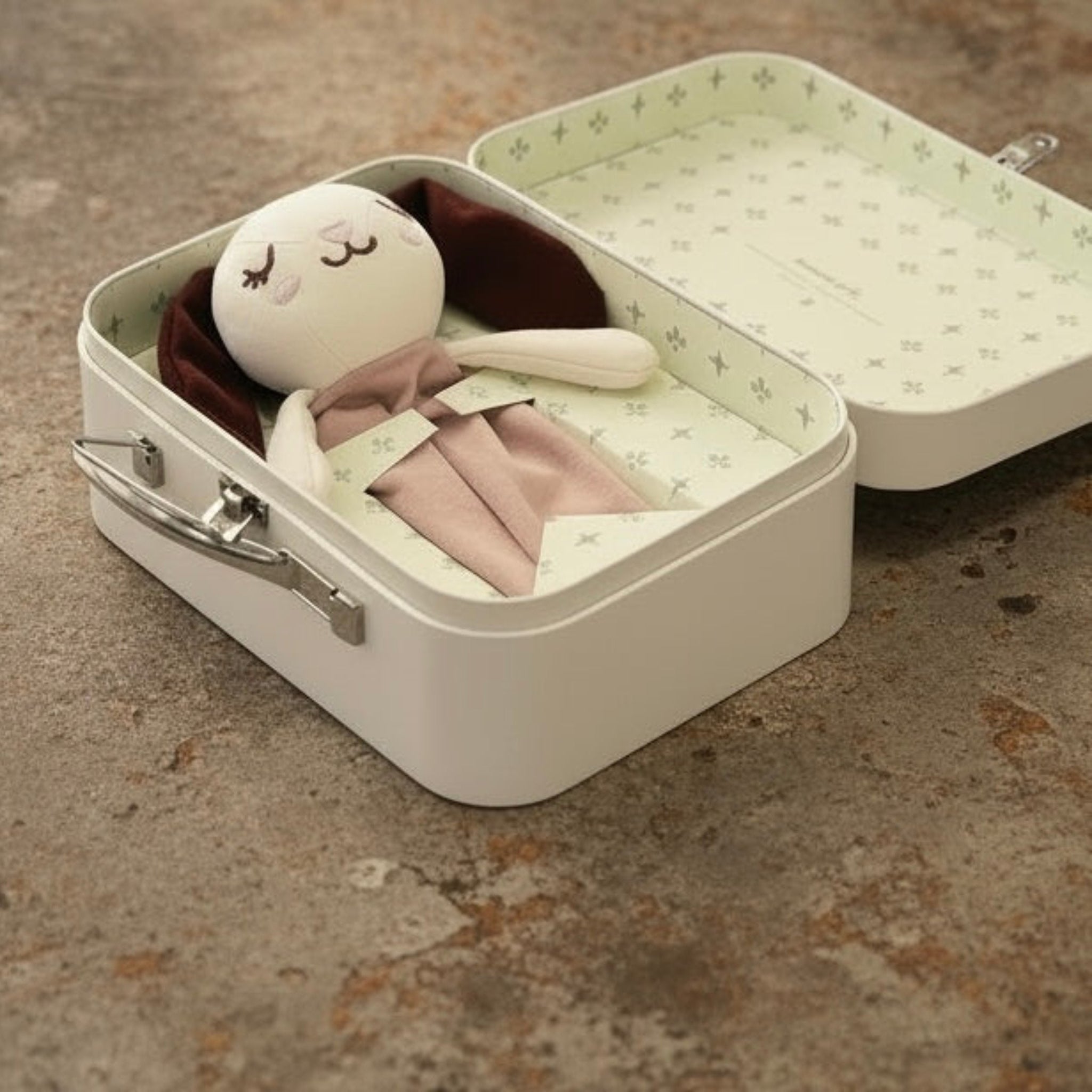 A small, sleepy softie keepsafe box opened on a textured concrete surface, containing a plush doll with a white face, dark brown hair, and pale pink clothing.