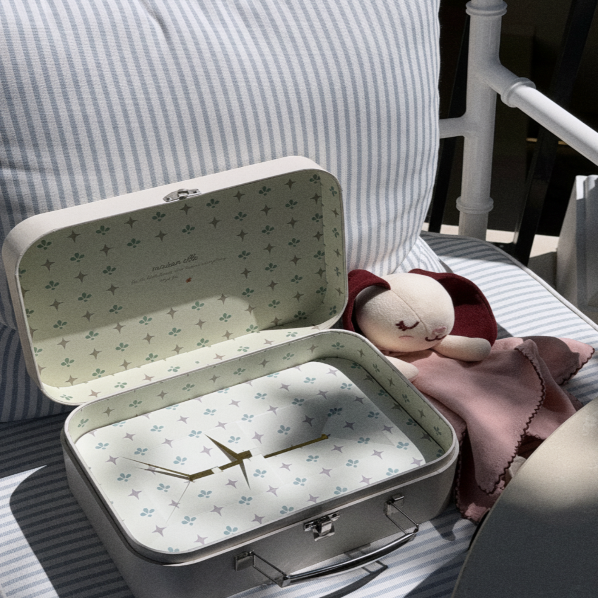 An open, sleepy softie keepsafe box clock with a floral patterned interior and gold hands, styled on a striped cushion next to a dusty pink sleepy softie.